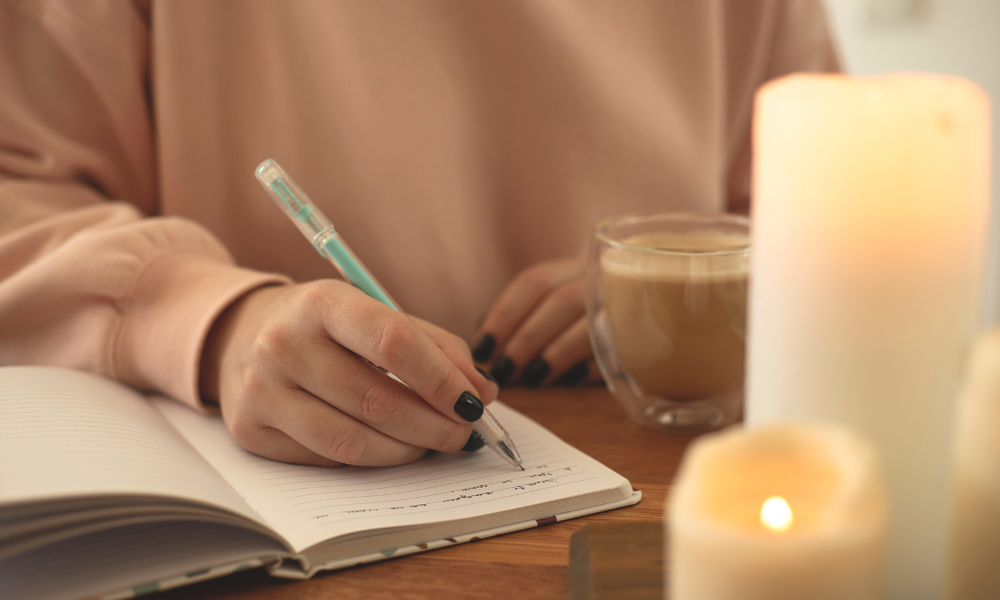 Woman writing in a journal with a coffee mug and candles.