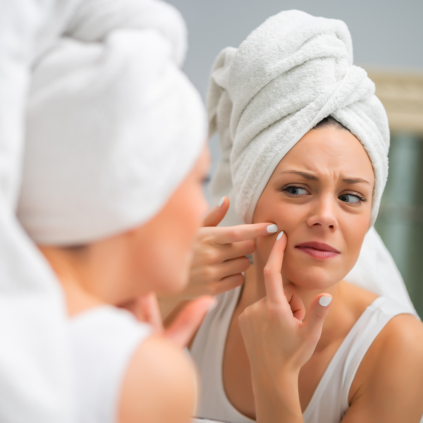 Woman with a towel on her head looking frustrated while examining her skin in the mirror and popping pimples.