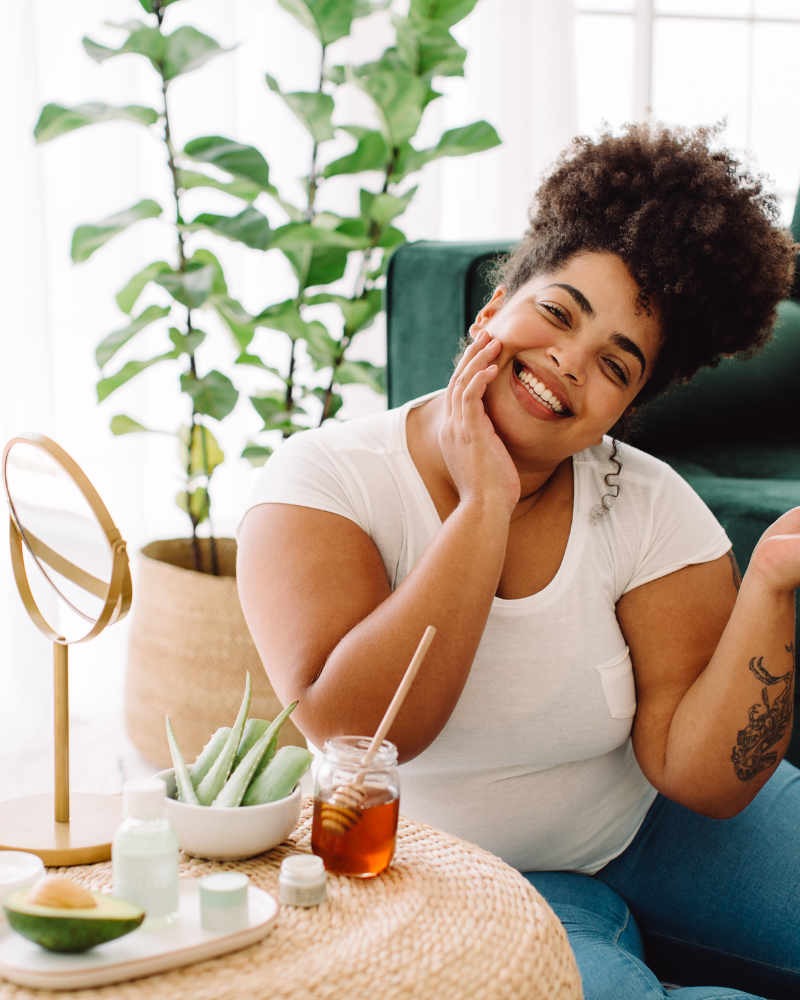 Woman smiling sitting next to a table with beauty, skincare, and self-care products.
