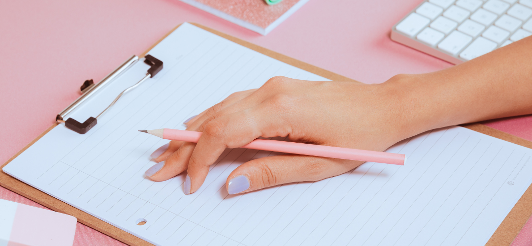 Woman holding a pen on a notepad with pink stationery and a pink background.